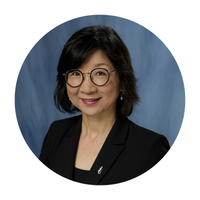 Portrait of Dr. Ji-Hyun Lee in front of a neutral blue background. She is smiling, sporting her black hair in neatly kept short feathered bob and wearing thinly rimmed black glasses.